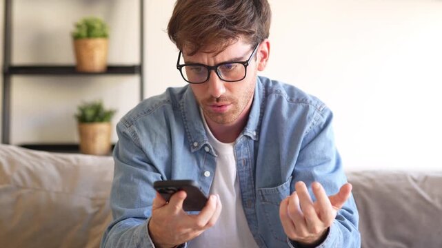 Angry young man sitting on sofa at home looking at smartphone with frustration, upset millennial experiencing internet or mobile problem, feeling stressed and annoyed by digital connection failure.
