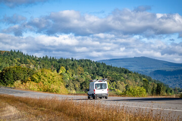 Small compact cargo mini van with ladders on the roof running on the autumn highway road with forest on the hills