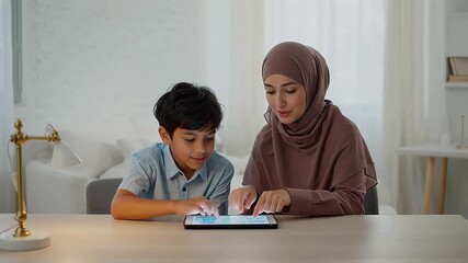 A woman and a young boy are sitting at a table, using a tablet together in a bright, modern room. - Powered by Adobe
