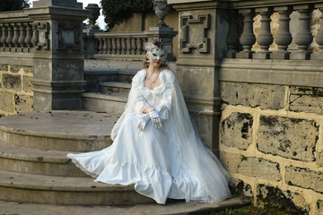 Fototapeta premium portrait of beautiful young blonde model wearing historical white wedding gown, fantasy cloak and butterfly masquerade mask. wandering ethereal figure in romantic fairytale castle garden background