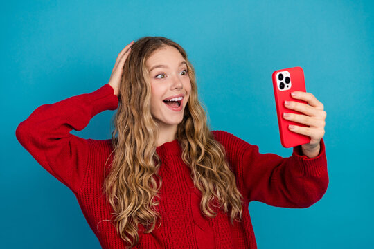 Cheerful woman in red sweater excitedly taking a selfie with a smartphone against a blue studio background