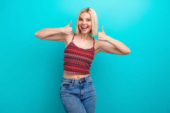Fototapeta Happy young woman giving thumbs up in a bright teal studio backdrop wearing a red patterned tank top and jeans
