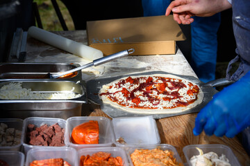 Chef placing fresh toppings on rolled out pizza dough on paddle to make individual wood fired pizza in food truck outdoors
