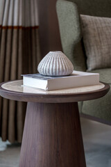 Home decor still life. A white textured ceramic vase and a book rest on a round wooden pedestal side table with a light marble top.
