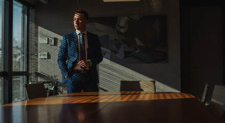 Confident businessman in sharp blue suit contemplates success in sunlit modern boardroom with striking abstract art