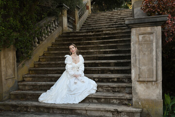 figure of female blonde model wearing white fantasy wedding gown. romantic sitting pose on staircase of historical fairytale castle location 
