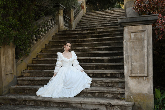 figure of female blonde model wearing white fantasy wedding gown. romantic sitting pose on staircase of historical fairytale castle location 
