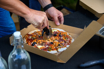 Man’s hands using a pizza cutter to slice a personal size wood fired pizza in a carboard box, cooked fresh outside in a food truck
