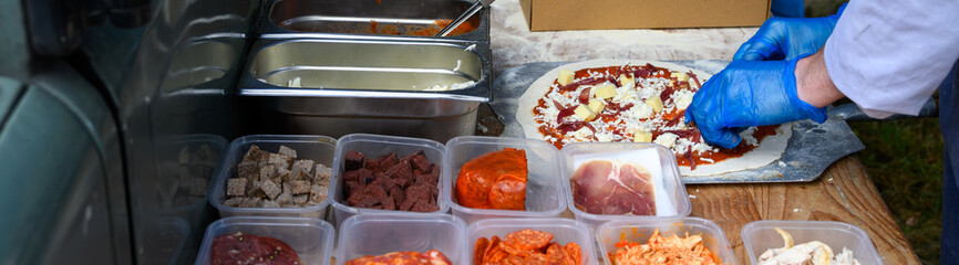 Chef placing fresh toppings on rolled out pizza dough on paddle to make individual wood fired pizza in food truck outdoors
