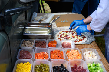 Chef placing fresh toppings on rolled out pizza dough on paddle to make individual wood fired pizza in food truck outdoors

