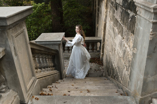 full length portrait of beautiful blonde female model wearing historical white gothic wedding gown, walking away back view, down staircase of romantic fairytale castle location with stone balconies