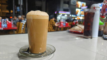 A glass of traditional teh tarik (pulled tea) with a distinctive layer of foam, served at a restaurant table in the evening. This warm beverage is popular in Southeast Asian eateries.