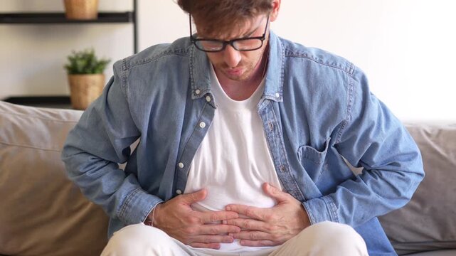 Young man sitting on sofa holding his stomach, feeling unwell and suffering from abdominal pain or digestive discomfort, showing sickness, nausea, and tension in a cozy home environment.
