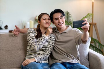 Young Asian couple take selfie together on sofa at home enjoying happy intimate moment share joyful lifestyle modern smartphone technology