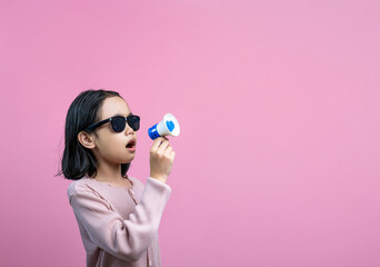 Cute young Asian girl wearing sunglasses holding a small megaphone and speaking against a pink...