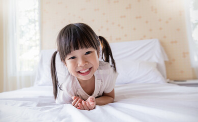 Portrait adorable funny little girl with pigtails smile brightly while lying on a white bed in a cozy cheerful home environment. Education preschool toddler.