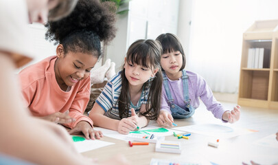 Group of happy, multicultural kids lying on floor and doing colorful drawings with crayons during creative indoor school activity.