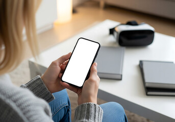 Woman Holding Smartphone with Blank Screen on White Table with Virtual Reality Headset and Books with Blonde Hair and Casual Clothing in Indoor Setting in Bright Ambient Lighting