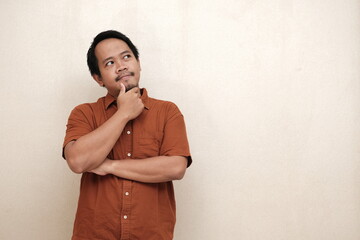 Southeast Asian man in a brown shirt standing with hand on chin, looking up thoughtfully as if...