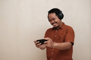 Southeast Asian man in a brown shirt smiling while playing a mobile game on his smartphone, wearing headphones in an indoor setting.
