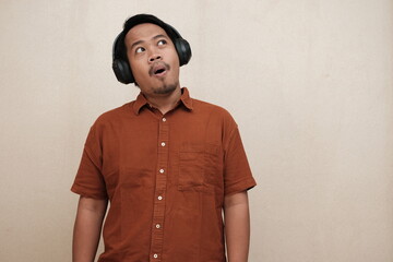 Southeast Asian man in a brown shirt looking joyful and surprised while listening to music on headphones in an indoor setting.