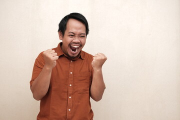 Southeast Asian man in a brown shirt clenching his fists and gritting his teeth, clearly showing...