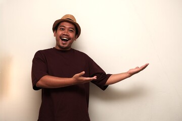 Excited Southeast Asian man in casual clothes and hat smiling while gesturing toward blank space,...