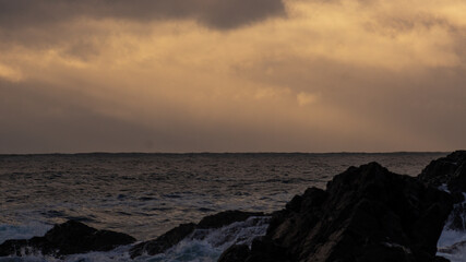 Rough ocean waters surge against jagged coastal rocks beneath an intensely cloudy, ochre-tinted sky at dawn or dusk.