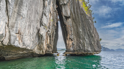 A sheer cliff rises in the ocean. A tourist canoe floats in the gap between the steep slopes. Mountains on the horizon. Blue sky, clouds. Philippines. Palawan. El NIdo