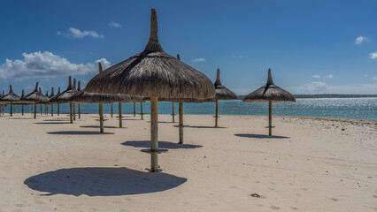 Rows of straw umbrellas on a sandy beach without people. Round shadows on white sand. A calm turquoise ocean. Blue sky, clouds. low season. Mauritius
