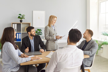 Positive businesswoman delivers presentation or meeting to team of colleagues in office, group work...