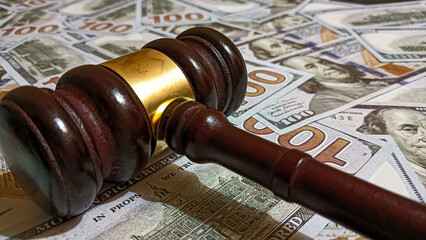 Wooden judge gavel placed on a pile of hundred-dollar bills representing law, justice, corruption, and financial judgment in legal or business contexts.