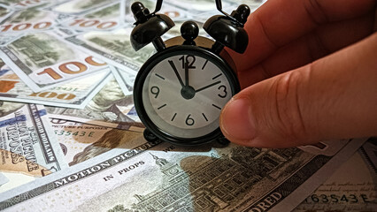 A black alarm clock placed on scattered US hundred-dollar bills, symbolizing the relationship between time and money, financial management, investment, and economic value.