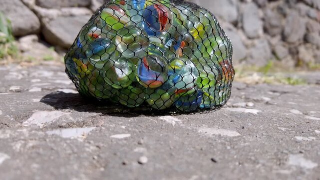 A mesh bag of vibrant marbles sits on sunlit stone pavement. The mix of colors in strong sunlight contrasts sharply with the textured, rustic, outdoor setting.