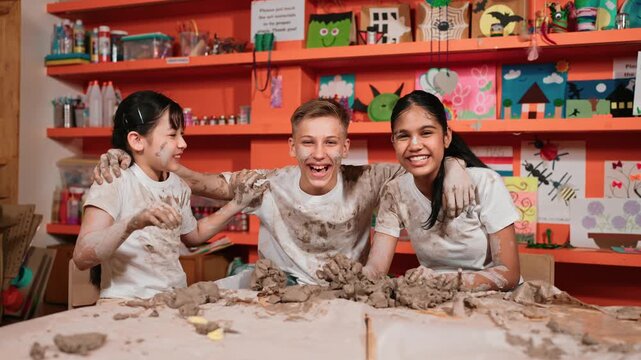 Smiling happy cute girl put piece of clay on smart caucasian boy face. high school student looking at camera while group of diverse children smile to camera at workshop. Creative activity. Edification
