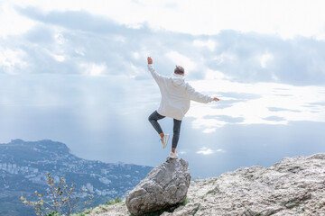 A man stands on one leg on a rock high in the mountains. A young woman in a white hoodie and black...