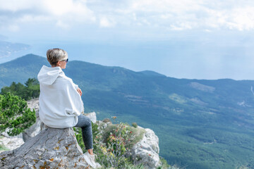 A woman in a white hoodie and sunglasses sits high on a mountain, looking down on the city and sea. A magnificent view of beautiful nature. Sports and activity.