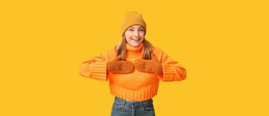 Mature woman in stylish sweater, hat and warm mittens on yellow background