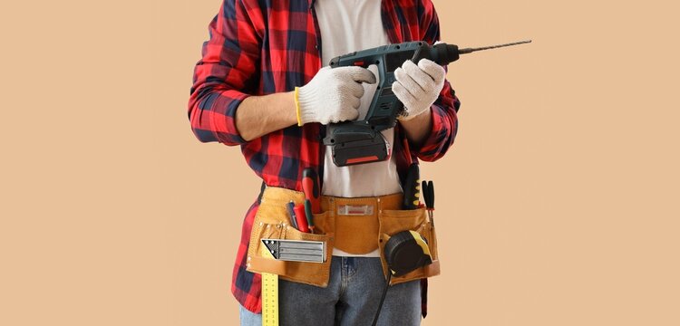 Male worker with drill and tools belt on beige background, closeup