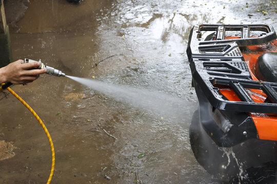 Person washing dirty garage floor with high pressure water hose and nozzle. Focused on cleaning concrete surface next to an all terrain vehicle after off road use