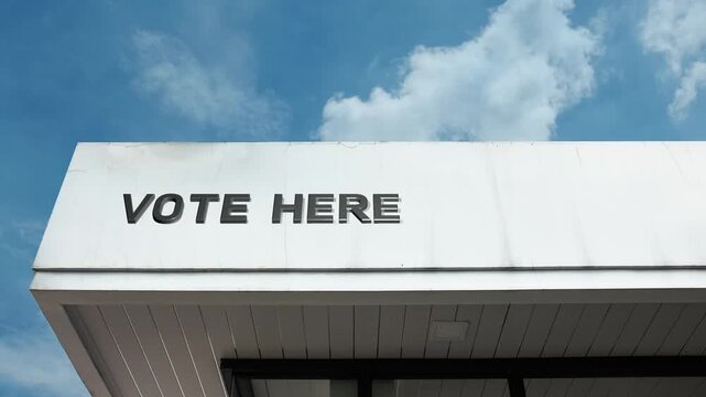 A "Vote Here" word sign displayed on a public or governmental building under a clear blue sky, symbolizing democracy, elections, civic duty, polling place, political participation, and public service