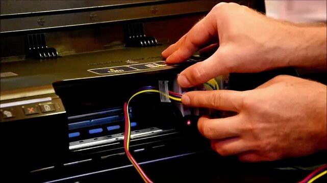 Hands carefully adjust the continuous ink supply system inside an open printer bay, aligning tubes and cartridges for smooth operation and uninterrupted ink flow