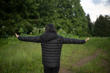 A girl with black hair and black clothes. The girl walks with her arms outstretched. Black clothes on the background of a green forest.