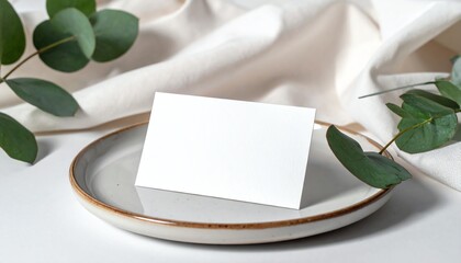 Blank card on ceramic plate, surrounded by green leaves and white silk, ideal for wedding invitations, branding, or elegant event stationery mockup.