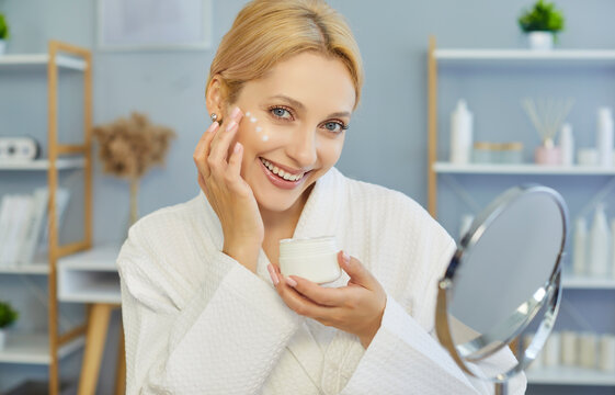 Fototapeta Beautiful smiling woman wearing bathrobe, smiling happily applying cream to her face and holding jar near mirror and bottles, self-care routine being enjoyed, contentment, personal well-being 