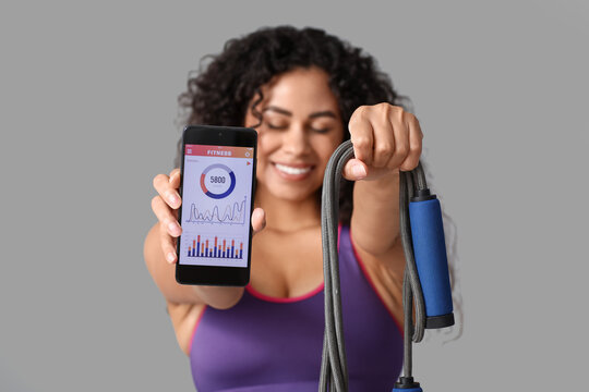 Sporty young African-American female coach holding mobile phone with calories counting app and jumping rope on white background. Concept of online training