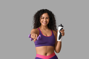 Sporty young African-American female coach with water bottle and dumbbells on white background
