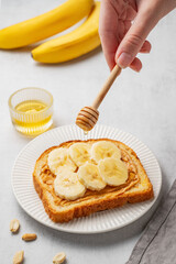 A hand holds a honey spoon over toast with peanut butter and banana on a white plate