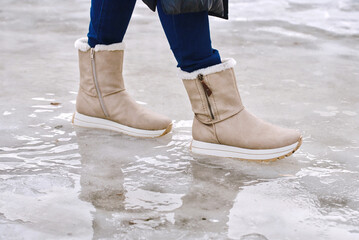 Woman stepping across slippery ice and frozen puddles outdoors in warm stylish beige winter boots with cozy fur lining, fashion comfort and safety during cold snowy weather. Slippery sidewalk