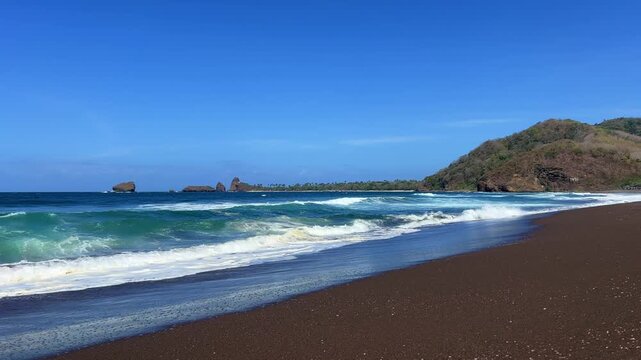 The sea in Jember, East Java. Indonesia. Watu Ulo beach with dark sand, mountains and coral rocks towering above the beach. The raging blue Indian ocean on a sunny day. 4К